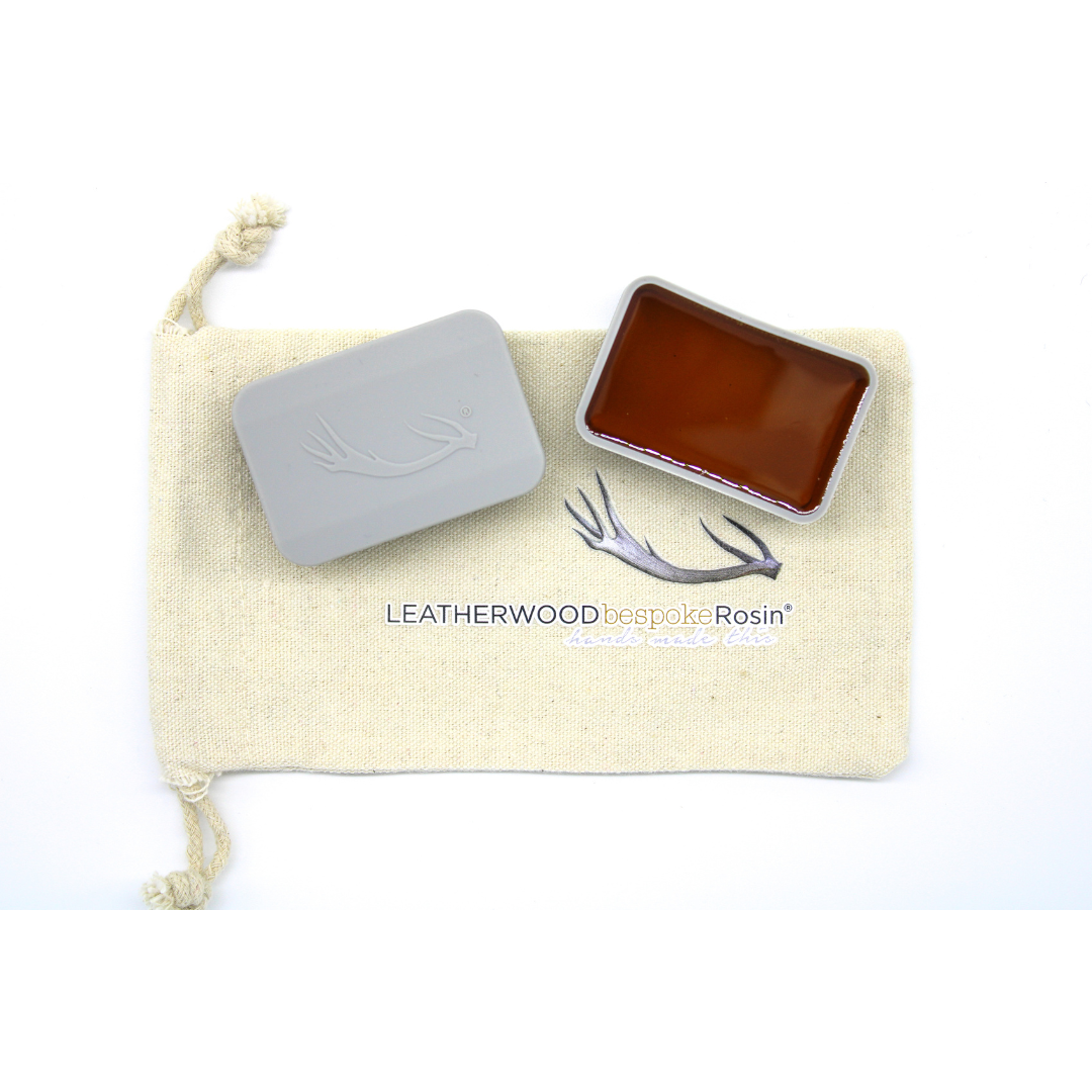 A gray rectangular container holding dark amber rosin rests open atop a light beige drawstring pouch.  The pouch shows the text: "LEATHERWOOD bespoke Rosin".