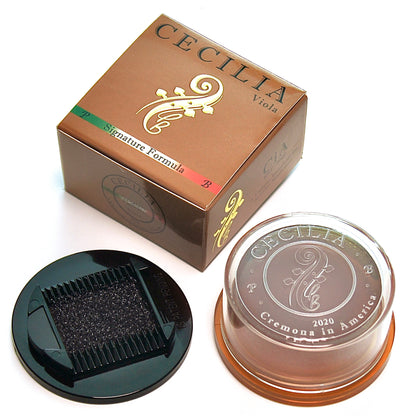 A rosin container sits next to its box.  The container is transparent, showing dark-brown rosin.  The box is brown with gold lettering: "CECILIA Viola Signature Formula."  The box and container feature a stylized viola design.